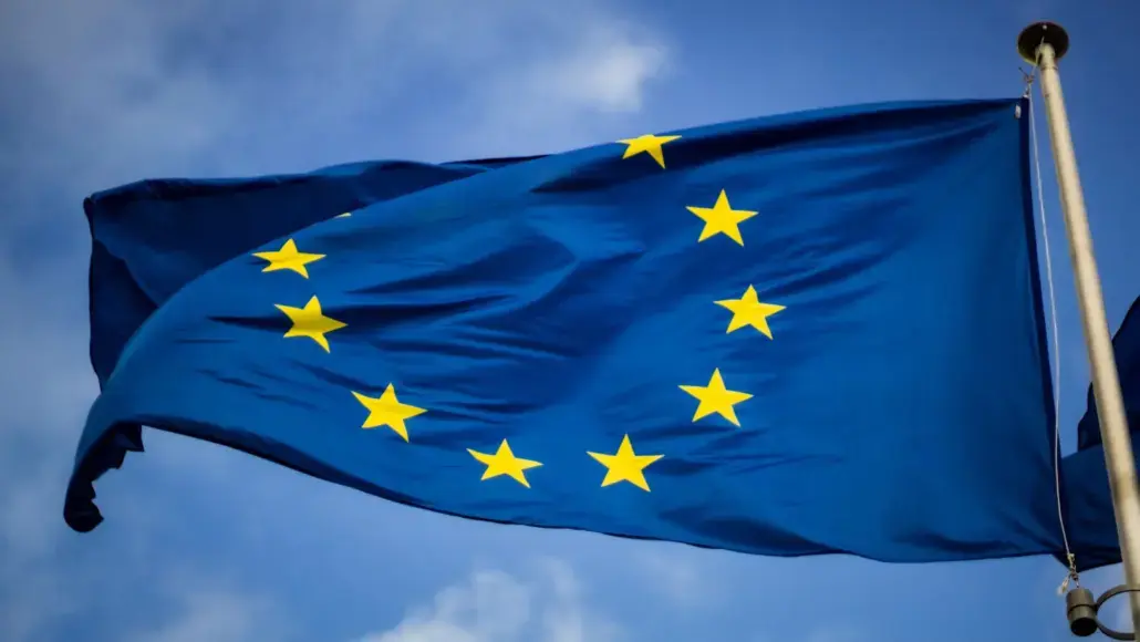 European Union flag waving on a flagpole with a circle of 12 yellow stars on blue field.