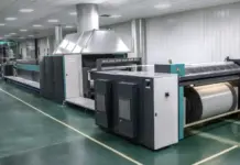 Karl Mayer Empowers AI Growth with Warp Preparation Technology Large industrial printing press in a clean factory, with roller drums and metal panels.