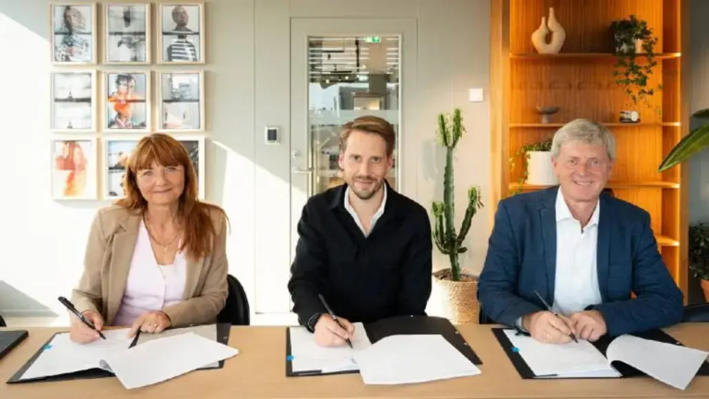 Three professionals sign documents at a conference table in a bright office setting.