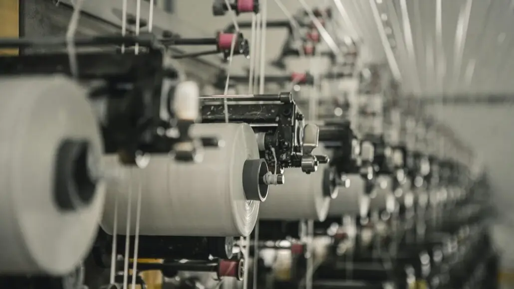 Close-up of a textile manufacturing loom with white yarn on large rollers and guides in a factory setting.