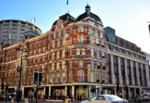 Frasers Group Completes Acquisition of Harvey Nichols from Administrators Harvey Nichols department store on a busy city street, red-brick facade and pedestrians nearby.
