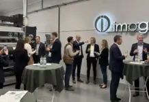 Imogo Unveils Digital Spray Dyeing Hub in Sweden for Sustainable Textile Production Professionals in business attire chat around high tables at an Imogo showroom with the company logo on the wall.