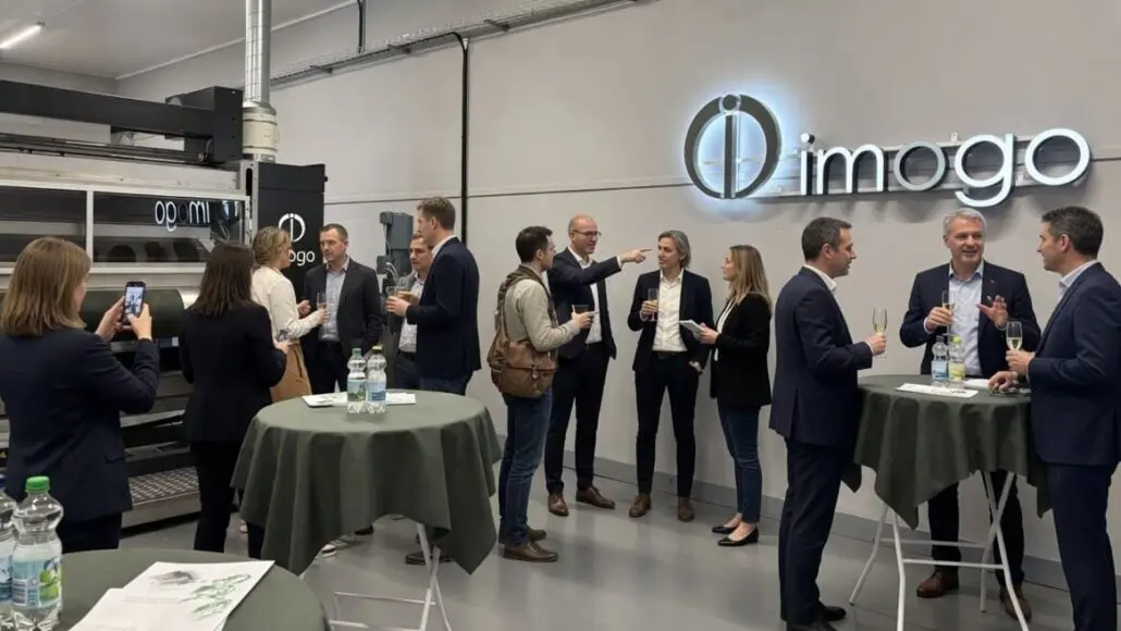 Professionals in business attire chat around high tables at an Imogo showroom with the company logo on the wall.