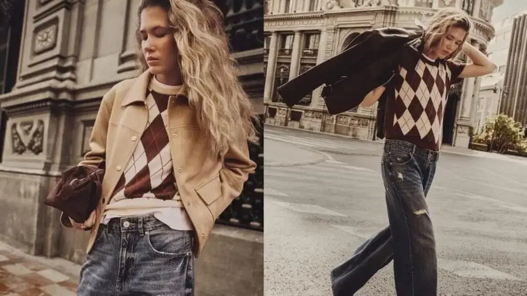 Young woman in argyle sweater and jeans poses near a building, wearing a tan jacket draped over her shoulders.