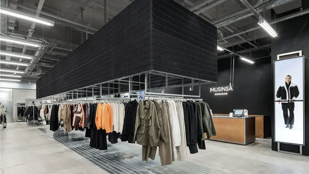 Clothing racks with coats in a modern fashion store; a wooden checkout desk and 'MUSINSA HONGDAE' sign in back.