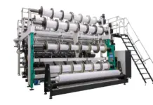 Karl Mayer Advances Footwear Production with New Double-Bar Raschel Technology Industrial fabric loom with multiple white spools and rollers, green frame, and metal access stairs.