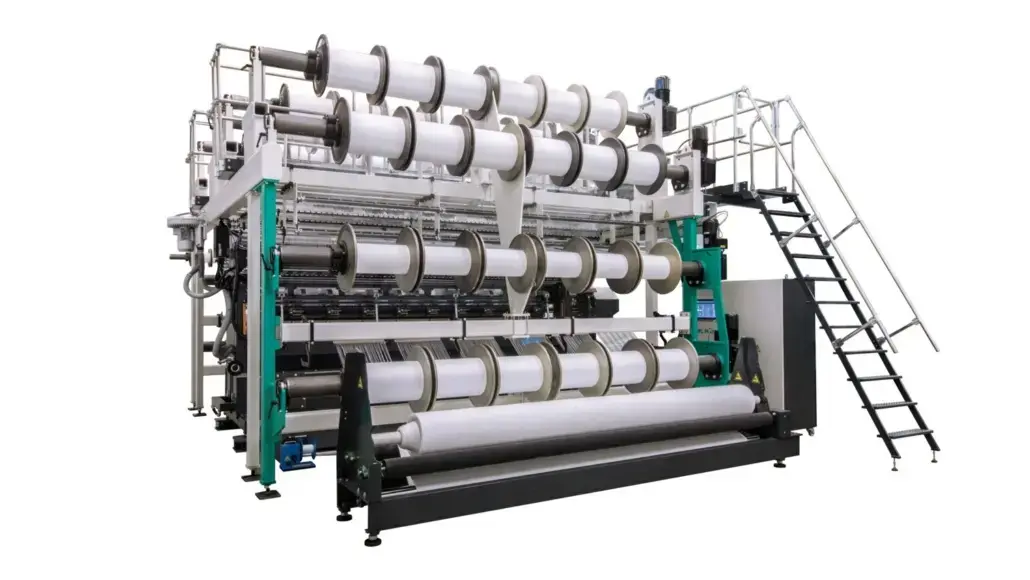 Industrial fabric loom with multiple white spools and rollers, green frame, and metal access stairs.