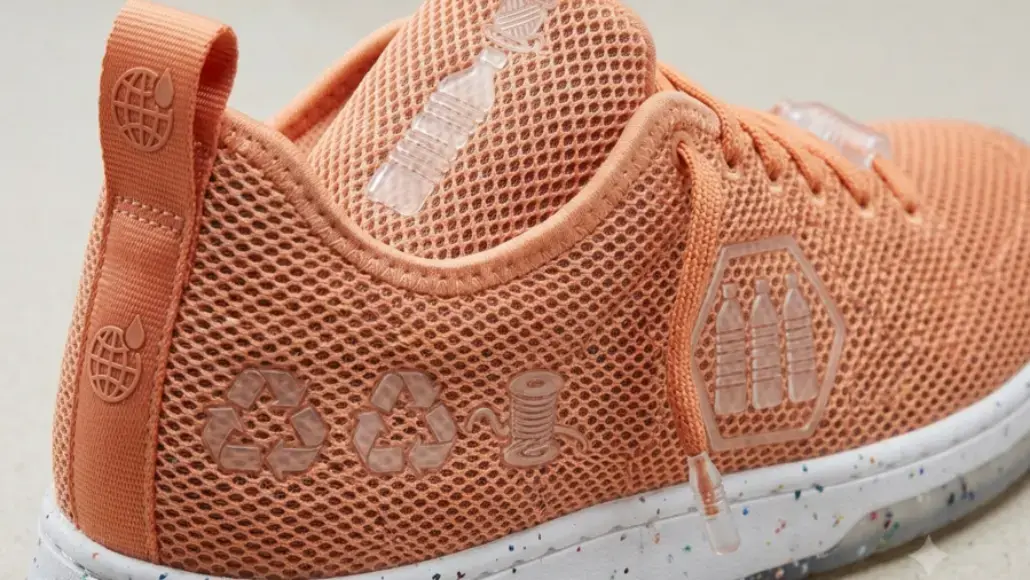 Close-up of a peach-colored mesh running shoe featuring recycled-material symbols and a speckled white sole on the heel side edge.