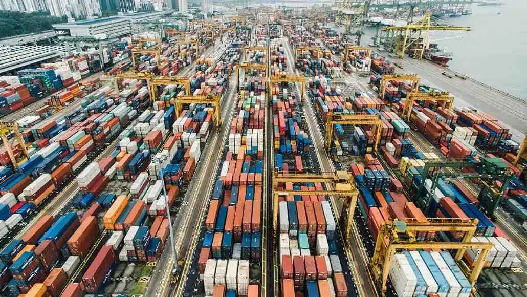 Aerial view of a shipping container yard with colorful containers and yellow gantry cranes at a busy port.