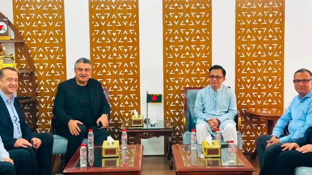 Four men in a formal meeting seated on sofas with glass tables and water bottles, in a room with decorative wooden partitions.