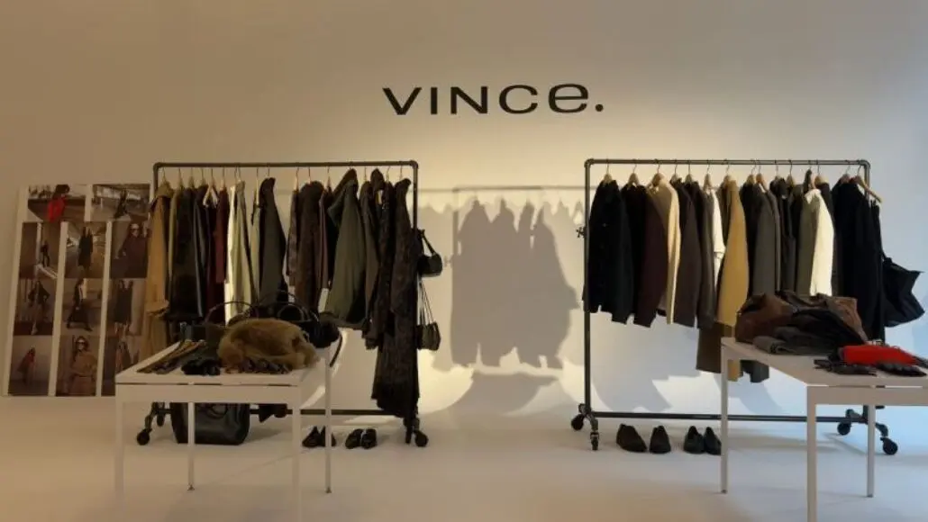 Interior of a Vince showroom with two clothing racks, tables, and coats on display under the Vince sign at the back.