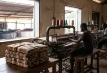 Advancing Local Value Retention in the African Textile Sector Weaving workshop with workers at looms, colorful thread cones overhead, and folded blankets stacked on a table nearby.
