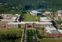 NC State University Selected to Lead New National Textile Manufacturing Initiative Aerial view of a university campus with red brick buildings, a central green quad, and surrounding trees; a lake in the distance.