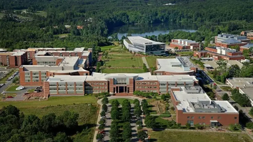 Aerial view of a university campus with red brick buildings, a central green quad, and surrounding trees; a lake in the distance.