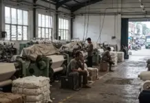 Indonesia’s Textile Industry Runs Far Below Capacity Amid Import Pressure Industrial textile factory interior with rows of weaving machines, wrapped fabric on each loom, and workers seated and sorting cloths under large windows.