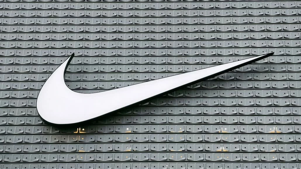 Large white Nike swoosh logo mounted on a gray tiled wall backdrop.