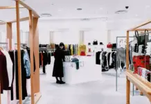 New Benchmarking Report Evaluates Trade Policy and Sourcing Challenges for 2026 Bright clothing store interior with wooden racks, hanging garments, and a white central display table; a shopper in a dark coat browses clothes.