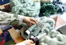 MELANGE Consortium Initiates Research into Circular Interior Textiles Hands handle chunky light-blue wool roving and spools of teal, mint, and gray thread on a table with fabric swatches, in a textile workspace.