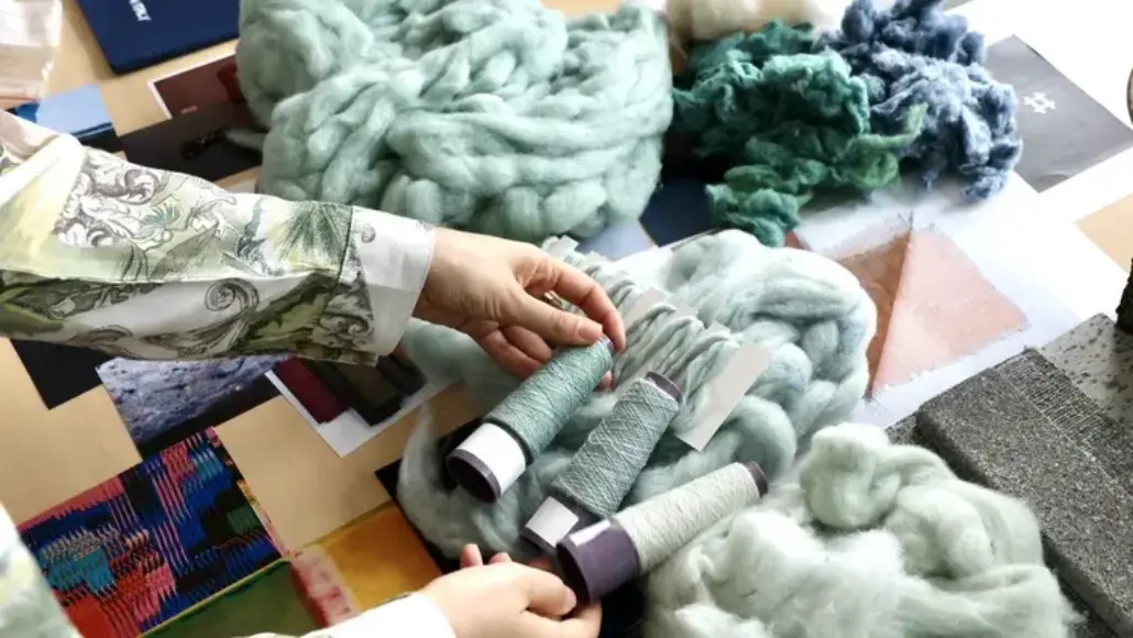 Hands handle chunky light-blue wool roving and spools of teal, mint, and gray thread on a table with fabric swatches, in a textile workspace.