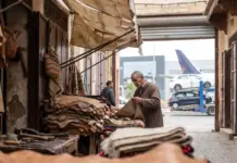 Morocco’s Textile and Leather Exports Drop 6.5% in H1 2026 Elderly man at a leather goods stall in a narrow market alley, sorting a leather hide among stacked hides and woven blankets in view.