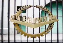 ADB and Modern Syntex Limited Sign $50 Million Loan to Expand Polyester Chips Production in Bangladesh Gold circular emblem with laurel leaves and a chevron reading 'Asian Development Bank' on a metal gate fence behind bars.