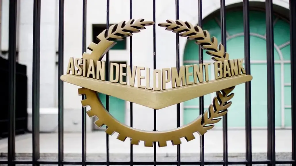 Gold circular emblem with laurel leaves and a chevron reading 'Asian Development Bank' on a metal gate fence behind bars.