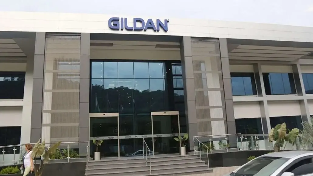 Front view of a modern Gildan office building with large glass doors, steps, and landscaped planters; a person walks by on the left.