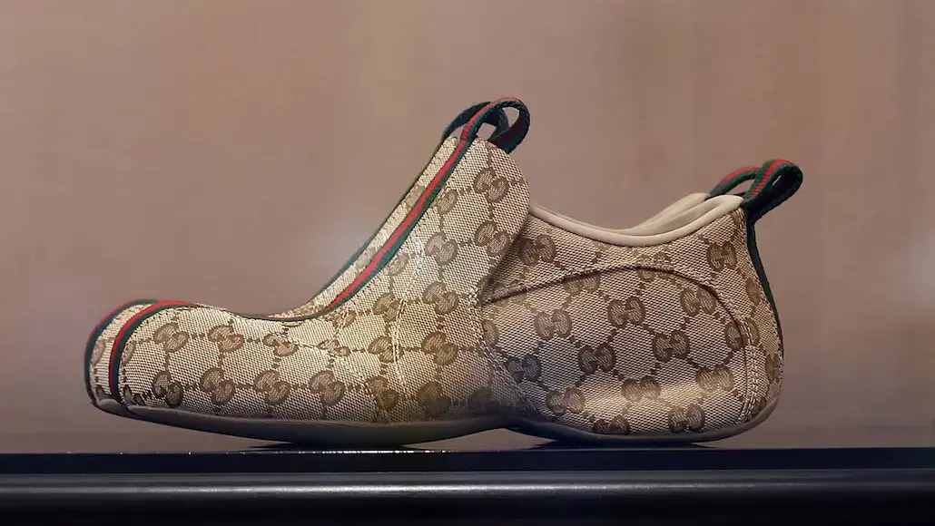 Beige Gucci monogram slip-on loafer with red‑green stripe and heel tabs on each side.