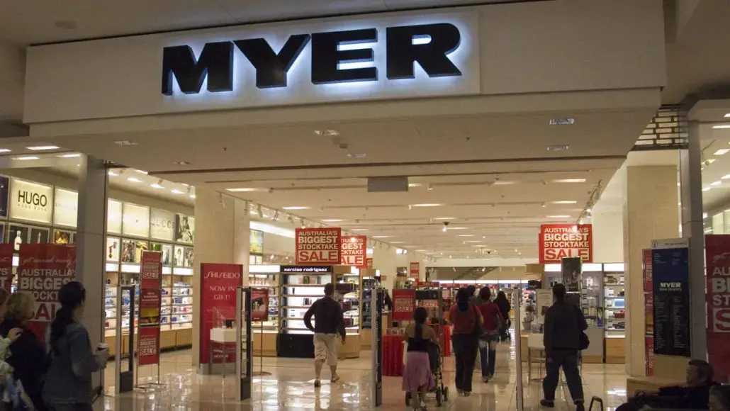 Entrance to a Myer department store with the large MYER sign overhead and shoppers walking through the aisles, red sale signs visible throughout the store.