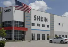 SHEIN Expands US Fashion Fulfilment With Indiana Hub SHEIN warehouse-style storefront with a waving American flag and '5400' on the facade, parked cars nearby.