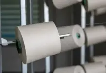 Cotton’s Share of Global Fiber Demand Falls to 21.1% as Synthetics Surge to 71.7% White yarn wound around a spool on a metal textile machine spindle, mid-rotation in a factory setting.