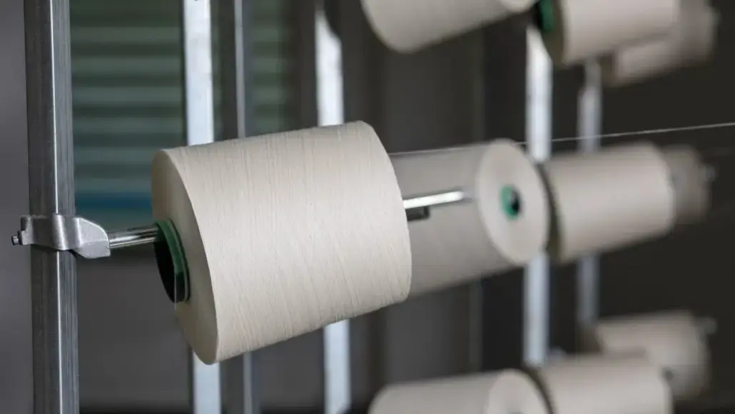 White yarn wound around a spool on a metal textile machine spindle, mid-rotation in a factory setting.