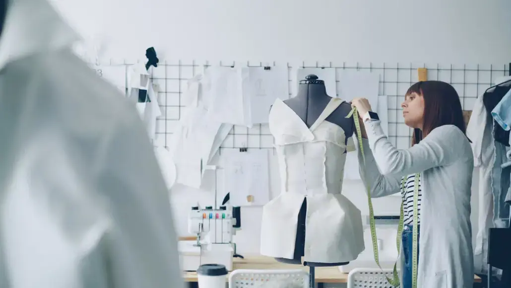 Designer in a studio takes measurements on a white dress form using a tape measure.