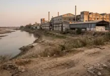How Water Scarcity Impacts Fashion Supply Chain Costs Industrial factory complex along a calm river at dusk, with a dry, cracked riverbank in the foreground and sparse vegetation.