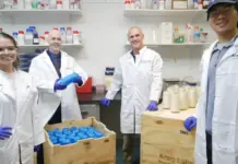 Kraig Biocraft Laboratories Ships First Commercial Spider Silk Order to Global Sports Brand Four scientists in white lab coats and blue gloves pose in a lab, with shelves of bottles behind and blue-capped containers in wooden crates front left and right.