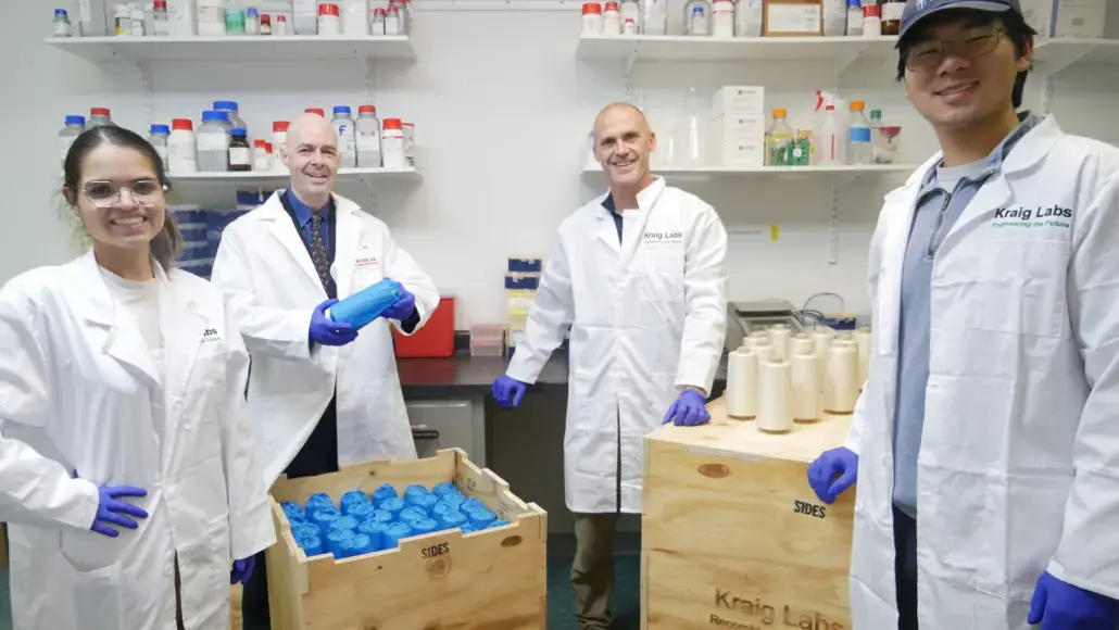 Four scientists in white lab coats and blue gloves pose in a lab, with shelves of bottles behind and blue-capped containers in wooden crates front left and right.
