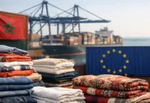 Morocco Textile Exports to EU Exceed €1 Billion in First Five Months of 2026 Stacks of folded blankets and textiles at a harbor, with shipping containers, cranes, and a cargo ship in the background; Moroccan flag visible on a container on the left.
