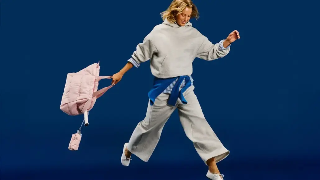 Woman in a gray hoodie and wide-leg pants leaps mid-air, pink backpack swinging behind her against a blue background.