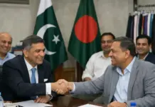 Pakistan and Bangladesh Textile Bodies Plan Joint Business Meetings and Exhibition Participation Two men in business attire shake hands across a conference table as colleagues smile in the background, with Pakistan and Bangladesh flags behind them.
