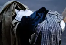 US Congressional Legislation Introduced to Combat Textile Waste and Promote Circularity Pile of clothes spilling out of a metal trash can on a sidewalk or street edge, including shirts and jackets in various colors (blue, gray, tan).