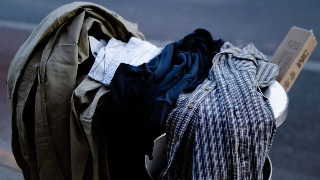 Pile of clothes spilling out of a metal trash can on a sidewalk or street edge, including shirts and jackets in various colors (blue, gray, tan).