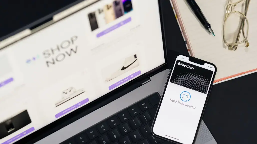 Laptop displaying an online shop with a prominent 'Shop Now' banner; a nearby iPhone shows Apple Pay Cash ready to pay