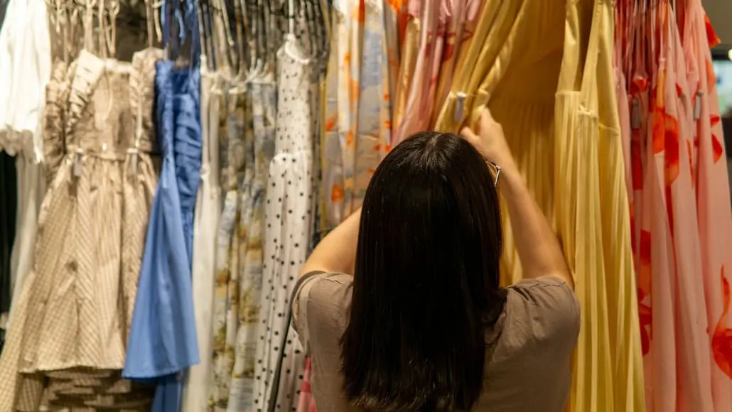 Back view of a person with dark hair in a beige top choosing a yellow dress from a rack of colorful dresses in a clothing store.