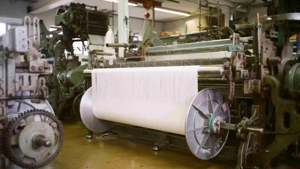 Industrial textile loom feeding a wide white fabric roll through machines in a factory setting.