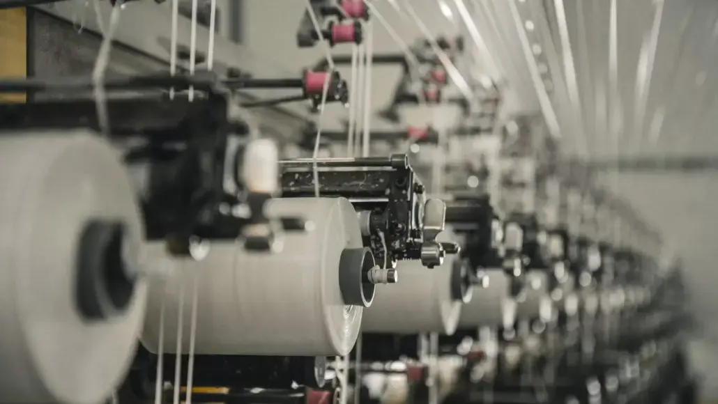 Row of textile manufacturing machines with white thread spools feeding along a production line in a factory setting