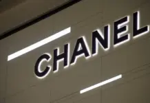Chanel Doubles Down on China as Luxury Rivals Close Stores CHANEL logo on a light tiled wall with bold black letters and subtle backlighting.