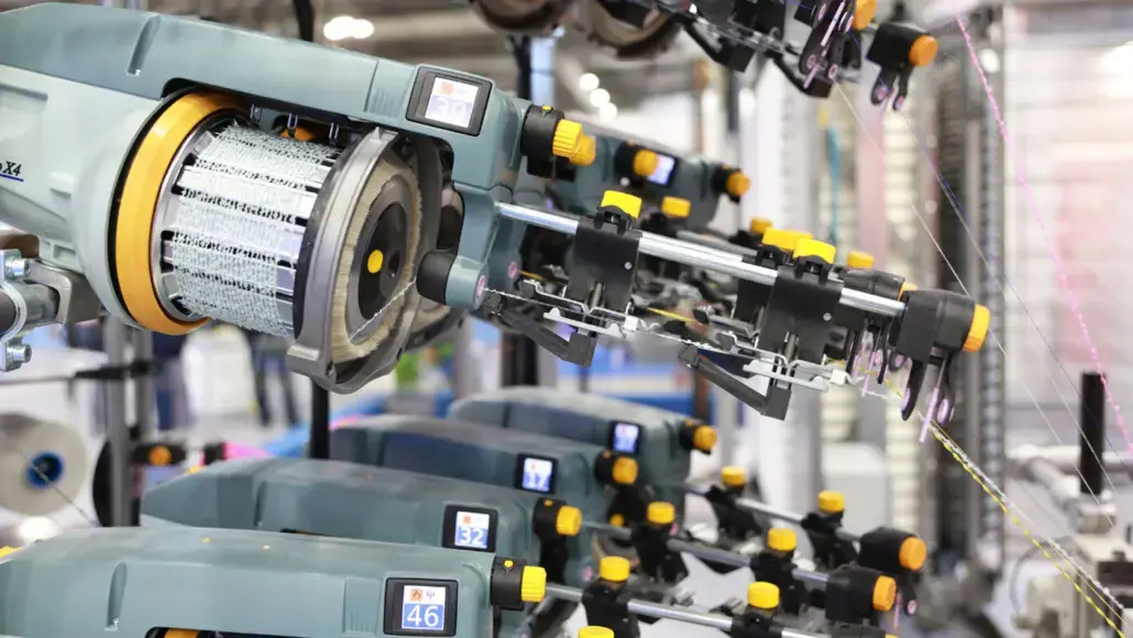 Close-up of an automated industrial knitting machine with multiple needle heads and yellow adjustment knobs in a manufacturing line.