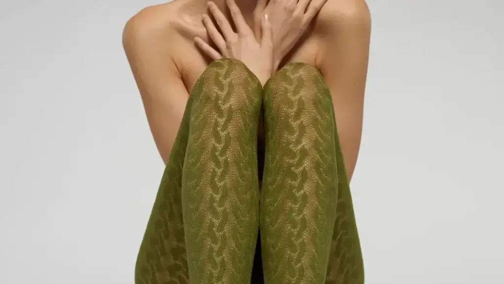 Person seated with knees drawn up, wearing olive-green patterned tights; arms cover their chest and shoulders are bare.