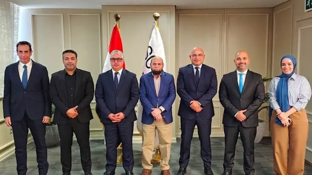 Seven professionals in suits pose for a group photo in a hallway with two flags behind them (red and white). They stand in a line, facing the camera, formal attire.