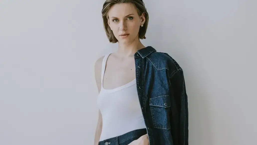 Woman posing in a white tank top with a denim jacket draped over one shoulder, against a light gray background.